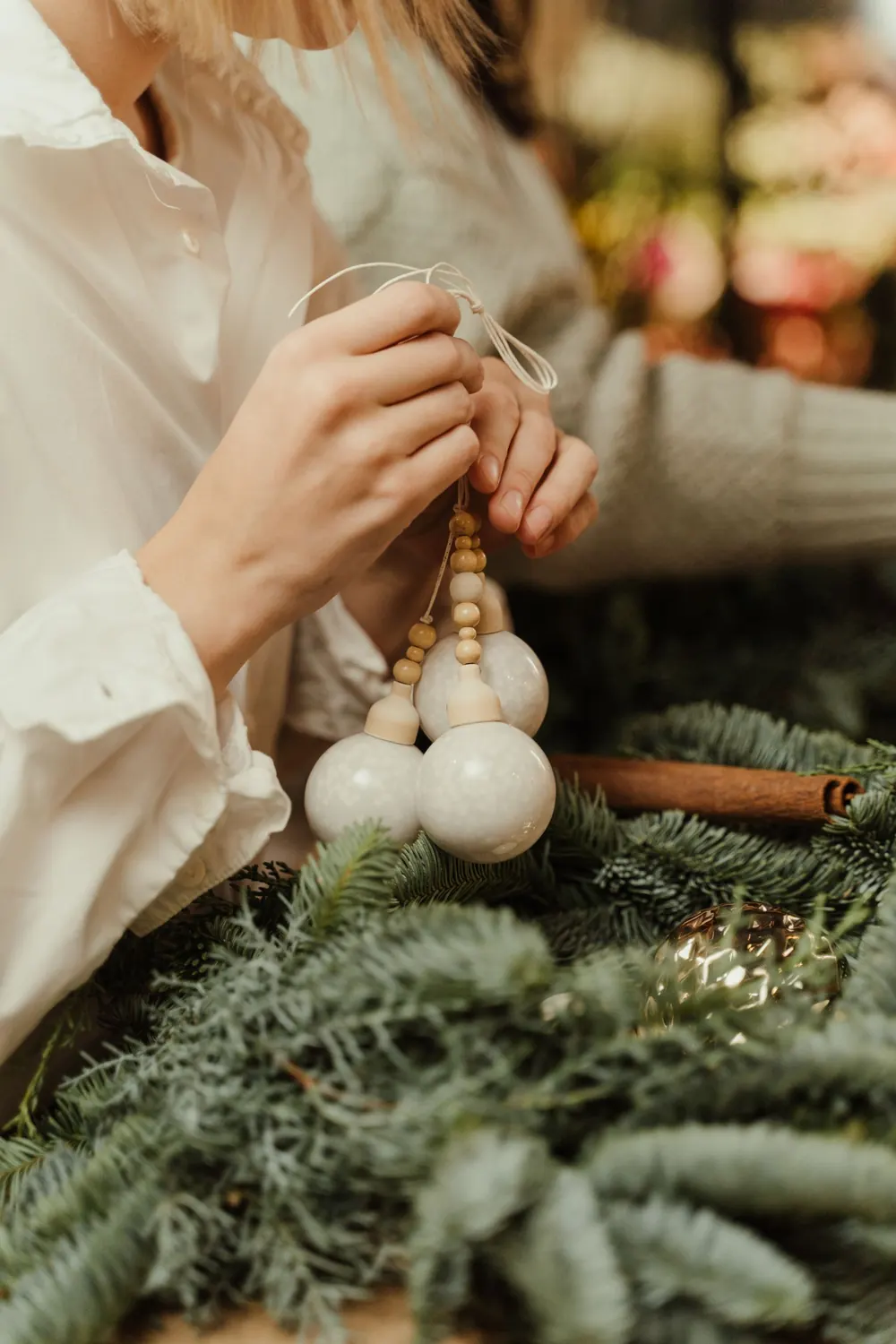 Woman holding white Christmas ornaments with fir leaves in background. Festive holiday preparation.